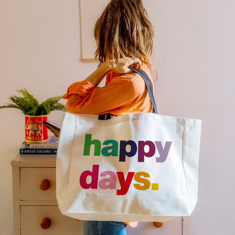 A white oversized slogan tote bag with contrasting black handles, printed with the phrase Happy Days in multi-coloured text